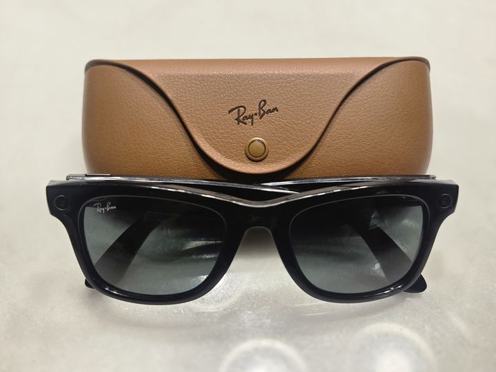 A pair of black camera smart glasses in front of their leather charging case